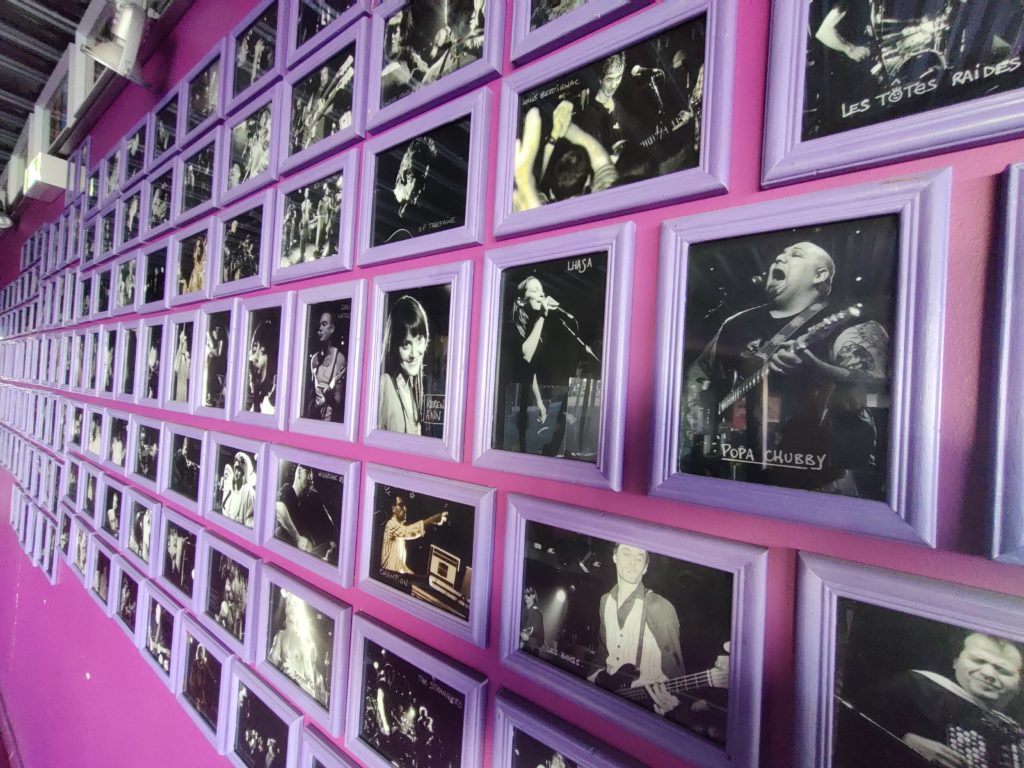 Photo représentant un mur tapissé de photos des groupes de musiques encadrés qui ce sont produit sur la scène de La Luciole.